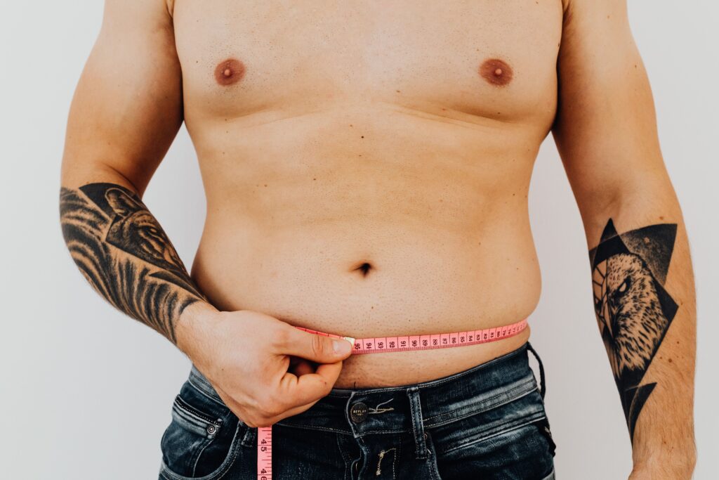 A tattooed male patient measuring his waistline, representing the growing demand for male liposuction and abdominal contouring procedures in Bangkok, Thailand.