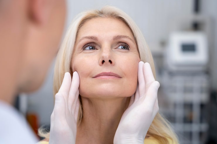 A surgeon in white latex gloves assessing a female patient's facial structure and jawline during a facelift consultation in Bangkok, Thailand.